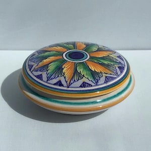 May include: A ceramic trinket box with a floral design on the lid. The lid features a central blue circle surrounded by orange, green, and blue leaf-like patterns. The box has a striped base with bands of orange, yellow, and teal.