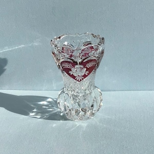 May include: A clear glass vase featuring a scalloped rim and a bulbous base with vertical fluting. The upper section is decorated with deep red heart-shaped panels, each containing an etched white floral design and intricate cross-hatch patterns.