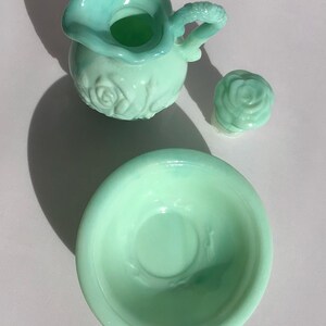K&ouml;nnte beinhalten: Ein hellgr&uuml;nes Glas-Set im Vintage-Stil, bestehend aus einem kleinen Krug mit Rosenmuster, einem rosenf&ouml;rmigen Deckel und einer kleinen Sch&uuml;ssel. Der Krug hat einen strukturierten Griff. Das Set wird auf einer hellen Oberfl&auml;che pr&auml;sentiert.