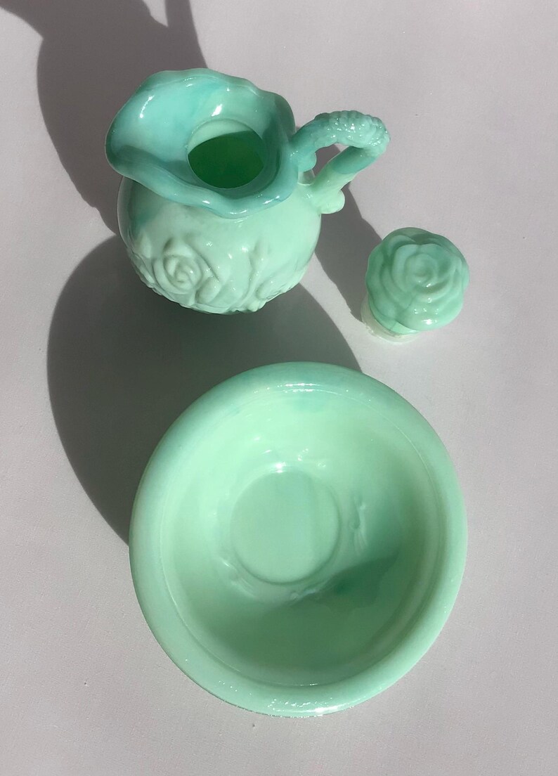 K&ouml;nnte beinhalten: Ein hellgr&uuml;nes Glas-Set im Vintage-Stil, bestehend aus einem kleinen Krug mit Rosenmuster, einem rosenf&ouml;rmigen Deckel und einer kleinen Sch&uuml;ssel. Der Krug hat einen strukturierten Griff. Das Set wird auf einer hellen Oberfl&auml;che pr&auml;sentiert.
