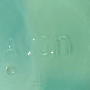 K&ouml;nnte beinhalten: Nahaufnahme einer hellt&uuml;rkisfarbenen Oberfl&auml;che mit dem gepr&auml;gten Wort "Avon". Die Buchstaben sind leicht erhaben und scheinen Teil eines gr&ouml;&szlig;eren Objekts zu sein. Die Oberfl&auml;che hat ein gl&auml;nzendes Finish.
