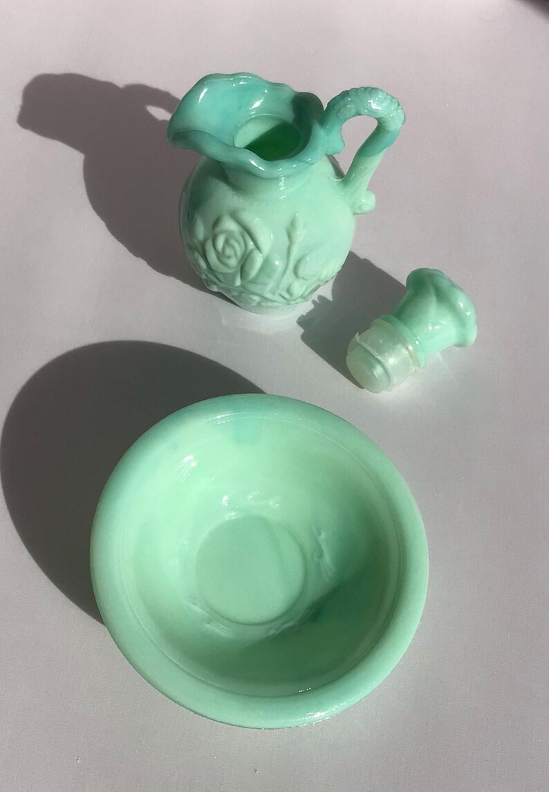 K&ouml;nnte beinhalten: Ein hellgr&uuml;nes Glas-Set im Vintage-Stil, bestehend aus einem kleinen Krug mit Henkel, einer Schale und einem Verschluss. Krug und Verschluss haben ein florales Rosendesign. Das Set ist wahrscheinlich f&uuml;r dekorative Zwecke bestimmt.