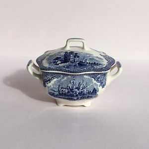 May include: A white porcelain sugar bowl with a blue and white floral design. The lid has a raised handle and features a scene of a castle.