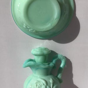 K&ouml;nnte beinhalten: Eine hellgr&uuml;ne Avon-Kanne und eine passende Schale. Die Kanne hat ein Rosenmuster und einen Griff. Die Schale ist rund mit einem floralen Muster. Das Avon-Logo ist auf der Unterseite der Schale sichtbar.