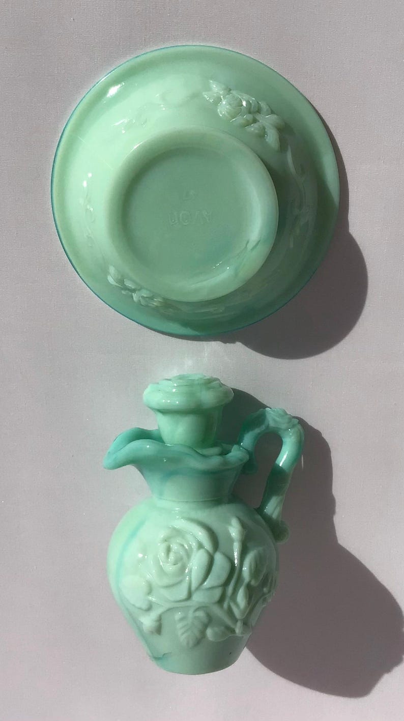K&ouml;nnte beinhalten: Eine hellgr&uuml;ne Avon-Kanne und eine passende Schale. Die Kanne hat ein Rosenmuster und einen Griff. Die Schale ist rund mit einem floralen Muster. Das Avon-Logo ist auf der Unterseite der Schale sichtbar.