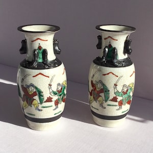 May include: Pair of white ceramic vases with black accents and painted figures in green, red, and yellow. The vases have a rounded body and a narrow neck.