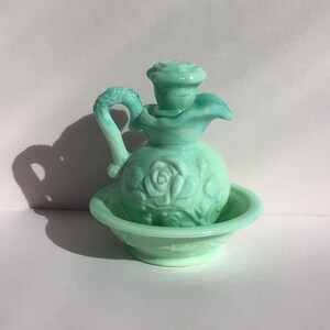 K&ouml;nnte beinhalten: Ein Vintage-Set aus hellgr&uuml;ner Glaskanne und -becken. Die Kanne hat ein Rosendesign und einen dekorativen Griff. Das Becken hat ein passendes Rosenmuster. Das Set wird auf einer wei&szlig;en Oberfl&auml;che pr&auml;sentiert.