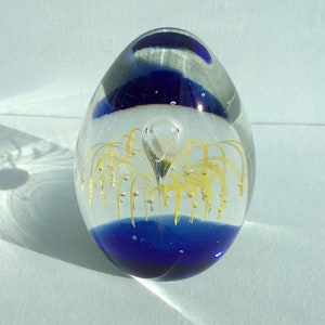 May include: A clear glass paperweight in the shape of an egg with a blue base and a yellow flower-like design inside.
