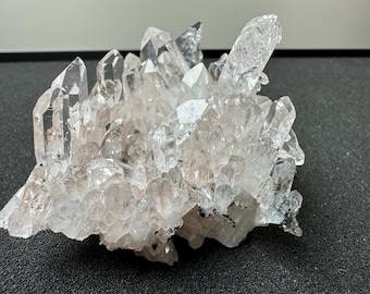 May include: A cluster of clear quartz crystals with a rough, natural texture. The crystals are arranged in a random, organic pattern, creating a unique and beautiful display.