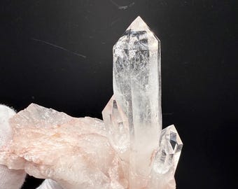 May include: A clear quartz crystal cluster with multiple points, set against a dark background. The quartz crystals are transparent and have a sharp, pointed shape. The base is a light pink color.