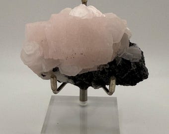 May include: A pink mineral specimen with a layered, textured appearance, mounted on a clear acrylic stand. The mineral has a soft, light pink hue with areas of white and is set against a dark, possibly black, base.