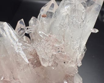 May include: A cluster of clear quartz crystals with sharp, pointed terminations. The crystals have a translucent quality, with some areas showing a hint of pinkish-red coloration. The arrangement creates a visually striking, natural formation.