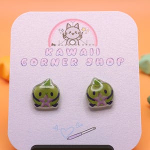 May include: A pair of green, rounded character stud earrings displayed on a white card labeled Kawaii Corner Shop. The earrings feature a small green creature holding a purple star. The card includes a line drawing of a pen and a heart.