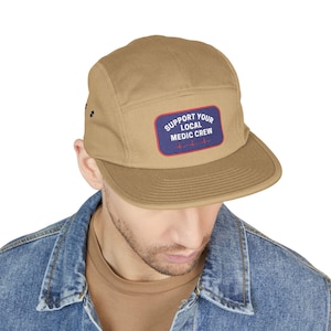 Peut inclure: Casquette cinq panneaux de couleur beige avec une visière plate. Le chapeau comporte un écusson rectangulaire avec le texte "SUPPORT YOUR LOCAL MEDIC CREW" en blanc sur fond bleu, avec un graphique de battement de cœur rouge.