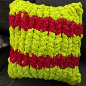 May include: A square, handmade pillow with a vibrant, textured design. The pillow features a bright lime green base with two bold, horizontal stripes of deep red. The plush, chunky knit construction adds to its cozy appeal.