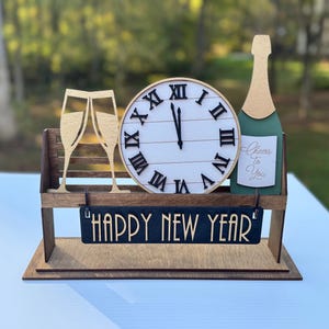 May include: Wooden New Year's Eve decoration featuring a clock, champagne bottle, and two champagne glasses. The clock shows midnight, and the bottle has a label that says "Cheers to You". A sign reads "HAPPY NEW YEAR".
