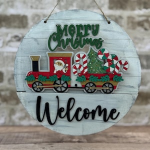 May include: Round wooden Christmas sign with a distressed white finish. Features a red train with Santa, candy canes, and a Christmas tree. The words "Merry Christmas" and "Welcome" are displayed.