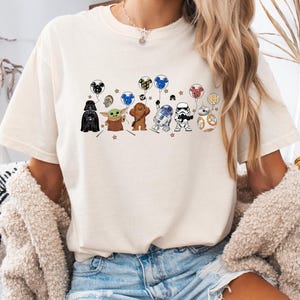 Peut inclure: T-shirt crème avec un motif de personnages de Star Wars et Disney tenant des ballons. Le design comprend Dark Vador, Grogu, Chewbacca, R2-D2, un Stormtrooper et BB-8. Les ballons ont des oreilles de Mickey Mouse.