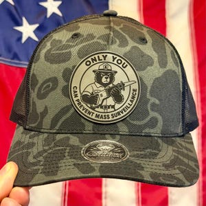 May include: A dark grey and black camouflage trucker hat with a black mesh back. A circular patch on the front features a bear wearing a hat and holding a reciprocating saw, with the text ONLY YOU CAN PREVENT MASS SURVEILLANCE around it.