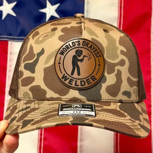 May include: A camouflage print trucker hat with a brown mesh back and a circular leather patch on the front. The patch features a black graphic of a welder and the text "WORLD'S OKAYEST WELDER". A black sticker on the brim reads "RICHARDSON 112".