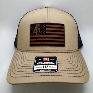 May include: A tan and black trucker hat with a brown leather patch featuring an American flag with the numbers "45 47" on it. The hat has a black mesh back and a brown leather label that reads "EST. 1970 RICHARDSON ORIGINAL 112 TRUCKER ADJUSTABLE".