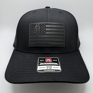 Puede incluir: Gorra de camionero negra con un parche de bandera estadounidense en blanco y negro con los números "45 47". La gorra es ajustable y tiene un logotipo de Richardson en la parte posterior.