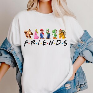 May include: White t-shirt featuring a graphic of several Mario characters, including Donkey Kong, Princess Peach, Mario, Luigi, Yoshi, and Bowser. The word "FRIENDS" is printed in black block letters below the characters.