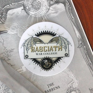 May include: A white circular sticker with a black and gold design. The sticker features a stylized image of a pair of wings with the text "Babciath War College" in a gothic font.