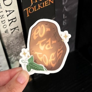 May include: A brown potato sticker with the words "Po-ta-toes" written on it in a stylized font. The sticker has a green leaf and a white flower on the bottom.