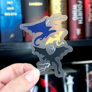 May include: A clear sticker with three dragons in silhouette. The dragons are blue, yellow, and black. The sticker has lightning bolts around the dragons.