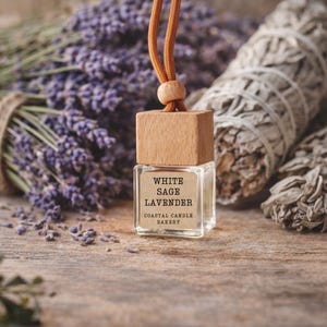 May include: A small, clear glass bottle with a wooden cap and a brown cord. The label reads "WHITE SAGE LAVENDER COASTAL CANDLE BAKERY." The bottle hangs in front of a bundle of purple lavender and white sage bundles, all on a wooden surface.