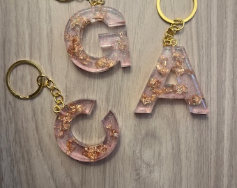 Op de afbeelding: Drie gouden sleutelhangers met roze harsletters, G, A en C, met gouden glitter ingebed in het hars.