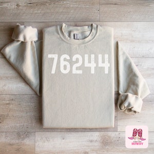 May include: A beige sweatshirt with the number "76244" printed in white across the front. The sleeves are rolled up. The sweatshirt is made of a soft material. The image includes a small logo with pink cowboy boots.