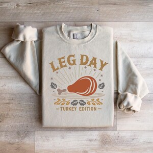 May include: A beige sweatshirt with the text "Leg Day - Turkey Edition" in brown and gold lettering. A stylised illustration of a turkey leg is featured in the centre of the design.