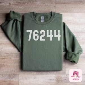 May include: A green sweatshirt with the number 76244 printed in white on the front. The sweatshirt is folded and laid flat on a wooden surface.