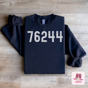 May include: A black sweatshirt with the number 76244 printed in white on the front. The sweatshirt is folded and laid out on a wooden surface.
