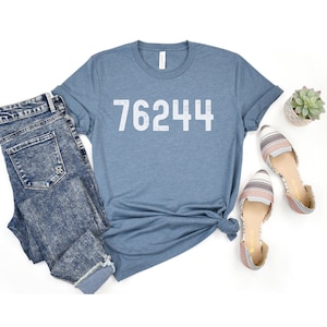 May include: A blue t-shirt with the number 76244 printed in white on the front. The shirt is laid out on a white background with a pair of patterned sandals and a potted succulent plant.