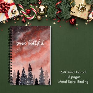 May include: A spiral-bound journal with a watercolour design featuring a red and orange sky, a forest silhouette, and the words "some bullshit." The journal is 15x20 cm with 118 lined pages and a metal spiral binding.