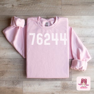 May include: A pink sweatshirt with the number 76244 printed in white on the front. The sweatshirt is folded in half and laid flat on a wooden surface. A small sticker with the text "Hometown Howdy" and a pair of pink cowboy boots is in the lower right corner of the image.