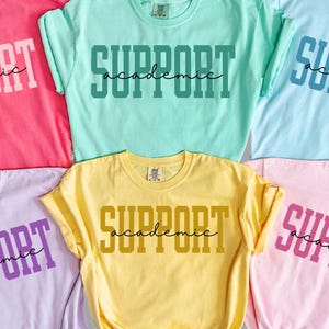 May include: Six colorful t-shirts in shades of pink, teal, blue, purple, yellow, and light pink. Each shirt has the words "SUPPORT ACADEMIC" printed on the front in a contrasting color. The shirts are displayed on a light background.