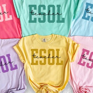 May include: Six colorful t-shirts in shades of pink, green, blue, purple, yellow, and light pink. Each shirt has the word "ESOL" in bold block letters and the word "teacher" in a cursive font.