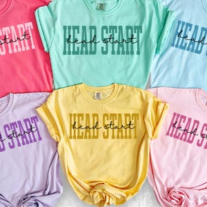 May include: Six colorful t-shirts in shades of pink, green, blue, purple, yellow, and light pink. Each shirt has the words "HEAD START" printed on the front in a contrasting color. The shirts are folded and displayed on a white surface.