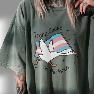 May include: Moss green t-shirt featuring a cartoon goose carrying a flag with the colors of the transgender pride flag. The text "Trans Goose on the loose" is printed above and below the goose illustration.