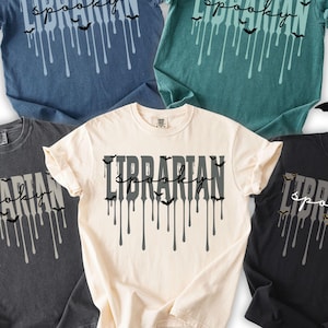 K&ouml;nnte beinhalten: F&uuml;nf kurz&auml;rmelige T-Shirts in Denim, Blaufichte, Pfeffer, Elfenbein und Schwarz. Jedes Shirt zeigt den Text "Librarian Spooky" mit tropfenden Linien und Fledermausgrafiken, perfekt f&uuml;r Halloween oder die Liebe eines Bibliothekars zur gruseligen Jahreszeit.