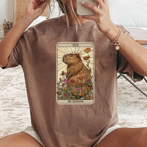 Pode incluir: Uma camiseta marrom com um gr&aacute;fico de uma carta de tar&ocirc; que apresenta uma capivara cercada de flores. A carta &eacute; rotulada como "XIX O Capibara".