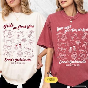 May include: Two t-shirts, one cream and one brick red, with white graphic designs. The cream shirt reads "Bride on Cloud Wine" and the red shirt says "Wine not? She's Tying the Knot." Both shirts have "Emma's Bachelorette" and "Napa Valley, CA, 2025" printed on them.