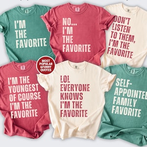 Könnte beinhalten: Sechs kurzärmelige T-Shirts mit Rundhalsausschnitt in Hellgrün, Karmesinrot und Elfenbein. Die Shirts zeigen abgenutzten weißen oder dunkelroten Text mit Sprüchen wie I'M THE FAVORITE, NO... I'M THE FAVORITE, DON'T LISTEN TO THEM, I'M THE FAVORITE, I'M THE YOUNGEST OF COURSE I'M THE FAVORITE, LOL EVERYONE KNOWS I'M THE FAVORITE und SELF-APPOINTED FAMILY FAVORITE.