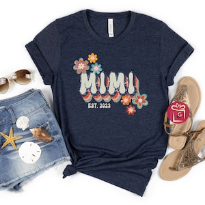 May include: A navy blue t-shirt with a retro-style graphic that says "MIMI EST. 2023" with colorful flowers.