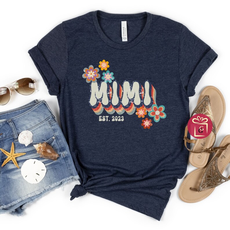 May include: A navy blue t-shirt with a retro-style graphic that says "MIMI EST. 2023" with colorful flowers.