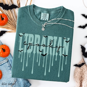 K&ouml;nnte beinhalten: T&uuml;rkisgr&uuml;nes T-Shirt mit den Worten "Librarian Spooky" in einer tropfenden Schriftart, verziert mit Fledermaus-Silhouetten. Eine silberne Halskette mit Sonnenanh&auml;nger liegt auf dem Shirt. Halloween-Dekoration.
